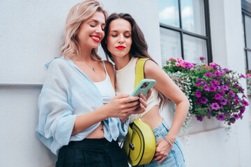 Two young beautiful smiling trendy female. Sexy women posing in street. Positive models having fun. Cheerful and happy. Hold smartphone, look at mobile cellphone screen, use phone apps, red lips