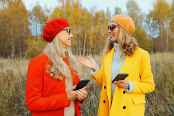 Wall Mural - Happy women friends talking with phone in autumn park, stylish girlfriends smiling holds smartphone