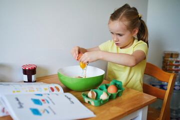 Wall Mural - Adorable preschooler girl cooking a dish on herself. Little child consulting with recipe book and breaking an egg into a bowl.