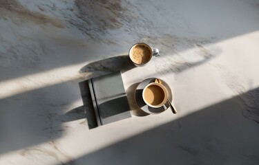 A marble surface with two cups of coffee and a small notebook in the sunlight.