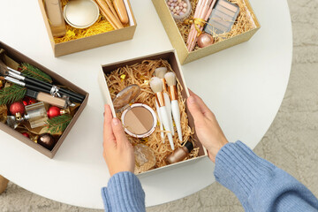 Canvas Print - Woman holding Christmas gift boxes with makeup brushes and cosmetics at home, above view