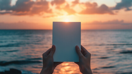 A person is holding a white book in front of a beautiful sunset