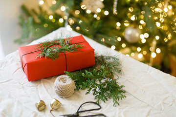  Zero waste Christmas concept. Stylish christmas gift and green branches on rustic table on background of christmas tree lights. Modern eco present wrapped in red paper with cedar wreath