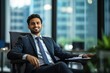 © Vadym - Handsome Indian businessman sits in office chair, wearing dark suit, blue tie. Smiling, looking directly at camera. Natural light fills office space, plant visible in background.