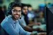 © Vadym - Man working in call center, wearing blue shirt and glasses. Seated at desk with computer monitor and headset. Busy office setting in background with people present.