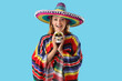 © Pixel-Shot - Pretty young woman in sombrero and colorful poncho holding decorative human skull on blue background