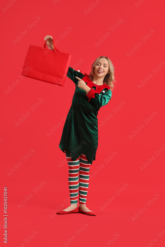 Young woman dressed as elf pointing at shopping bag on red background