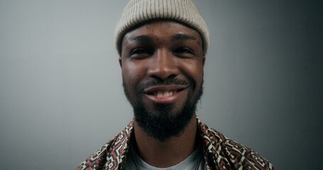 A bearded man in a hat, with an earring in his ear, smiling at the camera standing on a gray background in the studio, close-up of his face