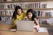 © fizkes - Positive young adult Indian students working on research study, educational project at laptop together, talking at computer with bookshelves in background, watching online content on screen