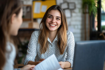 Two young women sit at a table with a laptop and papers, deep in discussion. A warm coffee shop ambiance surrounds them.