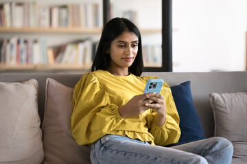 Wall Mural - Positive relaxed Indian student girl using smartphone in library. Young professional woman sitting on couch in office co-working lobby space, typing, chatting online on cellphone