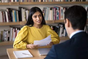 Sticker - Young Indian recruit manager woman talking to job candidate at interview, holding paper resume. Diverse student and teacher meeting in library, discussing research study