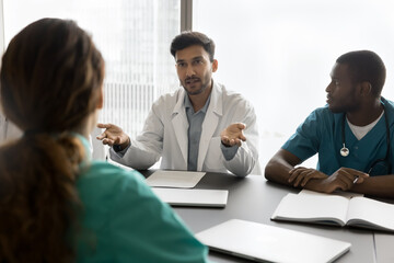 Young male doctor speak on multicultural team meeting ask advise in diagnostics choosing medical care tactics for severe patient case. Diverse medics colleagues meet for briefing at clinic staff room