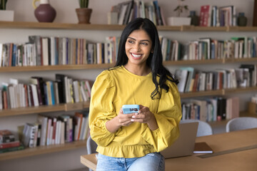 Wall Mural - Cheerful young Indian university student girl typing on cellphone, working on research study project in public library, looking away, smiling, thinking, leaning on work table in co-working