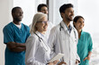 © fizkes - Inspired work group five diverse medics doctors stand together at hospital office look aside with candid smiles. Multiethnic medical service team with older leader motivated to help consult patients