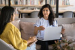 © fizkes - Positive engaged young Indian project manager talking to female colleague in co-working lobby space for meeting, holding laptop on lap, speaking, smiling. Two young co-workers discussing startup