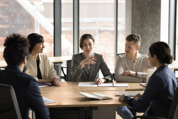 Canvas Print - Confident mentor woman training group of interns. Female boss instructing team of employees at meeting table, speaking, smiling. Diverse business partners negotiating on startup project