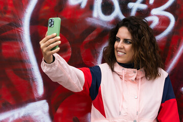 A young brunette in her 30s smiles as she takes a selfie with a wall covered in red graffiti in the background and wearing a pink jacket