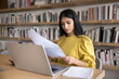 © fizkes - Serious Indian student working on research paper in library, reading essay, document. Focused young business woman reading investment agreement terms at laptop, financial reviewing reports