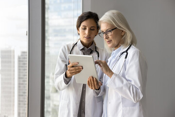 Young female doctor intern meet older age colleague ask question about patient history case show labs radiology results on tablet screen. Mature woman medic consult younger coworker at clinic corridor