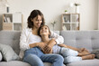 © fizkes - Tender moments. Smiling little girl clings closer to beloved single mom sitting on couch feeling safe protected in caring parent hugs speak talk to understanding mommy joking sharing childhood secrets