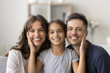 Happy loving family. Headshot portrait cheerful smiling parents kid hug on couch at home. Adorable little girl daughter tender caress faces touch cheeks of beloved millennial mom dad looking at camera