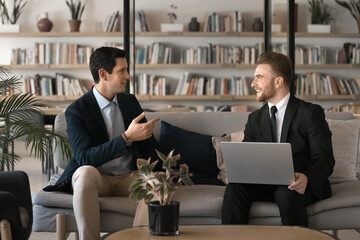 Poster - Positive successful young male business colleagues talking in co-working lobby space, siting on couch with laptop, discussing online project, job case, teamwork strategy, speaking, smiling