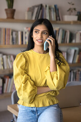 Wall Mural - Thoughtful positive young Indian student woman speaking on mobile phone in college library, making telephone call, enjoying communication, looking away, smiling. Casual vertical portrait