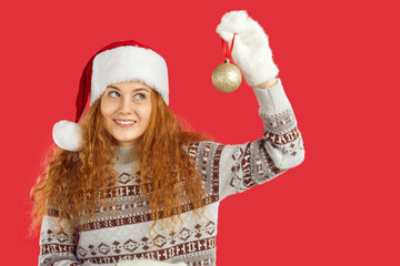 Christmas atmosphere. Joyful young woman in Christmas outfit is looking at Christmas tree ball in her hand. Redhead curly girl in santa hat, christmas sweater and mittens isolated on red background.