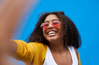 © Sanc/peopleimages.com - Portrait, happy and woman with selfie in studio for photography, heart glasses and social media post. Smile, girl and love sunglasses of profile picture, blog update and influencer on blue background