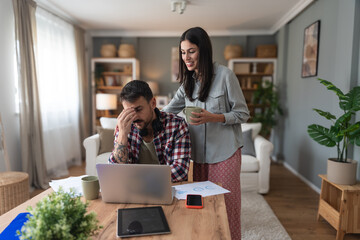 Young supportive wife brought a cup of coffee in morning to her tired husband who worked whole night on laptop computer. Freelance business man work online at home woman giving him support with coffee