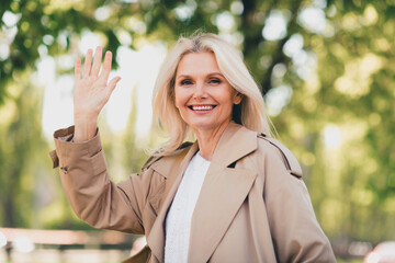 Portrait of positive happy retired woman smile spring good mood weekend wave hand hi wear beige coat enjoy outdoors outside park street