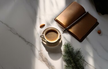 A cup of coffee, a closed notebook with a pen, and a pine sprig rest on a white marble surface with soft natural lighting.