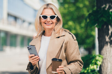 Wall Mural - Portrait of cheerful happy retired woman walking smile good mood hold device coffee weekend wear beige coat outdoors outside park street