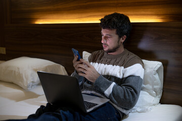 Happy young entrepreneur man on business trip works in the hotel room with his laptop and phone or smartphone