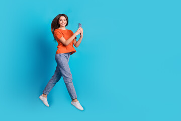 Sticker - Full size photo of pretty teen girl jump walk hold gadget wear trendy orange outfit isolated on blue color background