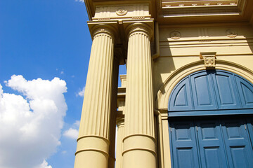 Close-up of classical architectural columns and arched doorway against a bright blue sky with fluffy clouds, showcasing historical elegance.