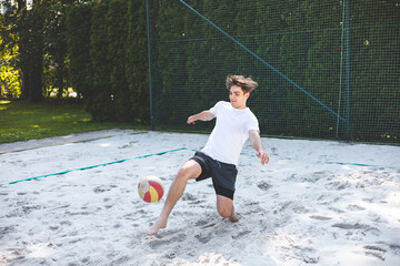 young man quickly reacts to a volleyball play by using his foot to keep the ball in play on a sandy court. His dynamic movement is captured mid-action, surrounded by a lush outdoor setting