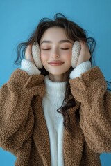 Wall Mural - Young woman smiles while wearing warm ear muffs and a cozy sweater against a blue backdrop