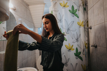 A young woman stands in a bathroom drying her hands with a towel, creating a calm and routine moment. The bathroom decor includes a floral-patterned shower curtain and clean, tiled walls.