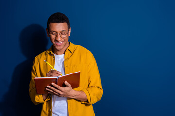 Poster - Photo of young smart guy in yellow shirt wearing eyeglasses holding copybook writing notes isolated on dark blue color background