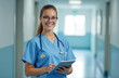 © Anton - A female doctor in glasses is wearing a medical suit, a stethoscope around her neck, tablet in hands, standing in the hospital corridor, looking at the camera and smiling