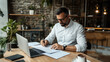 © altitudevisual - Man in white shirt working at desk with laptop and documents in modern office setting, surrounded by plants and brick wall background.