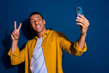 Poster - Photo of young happy guy wear yellow stylish shirt friendly showing v sign making selfie video isolated on dark blue color background