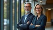 © Vilaphon  - Two confident executive managers, a mature business man and woman in their 50s, standing with arms crossed, smiling at the camera in a modern office environment with glass windows in the background