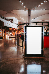 Wall Mural - A blank, vertical advertisement display screen mockup stands in a spacious, modern shopping mall or an airport terminal The sleek, glossy interior features stylish wooden elements and ambient lighting