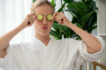 Woman holding slices of fresh cucumber and wearing bathrobe enjoying luxurious facial skincare spa in resort or hotel. Skin treatment for face and beauty care. Quiescent