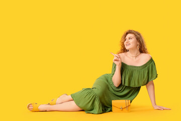 Wall Mural - Beautiful young woman in stylish green dress with chain bag sitting against yellow background