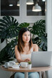 © Satori Studio - A young woman sits at a round table in a modern cafe, working on her laptop. The background features large green plants and natural light.
