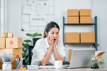 A young woman looks surprised while working on her laptop in a home office setting, surrounded by shipping boxes and plants.